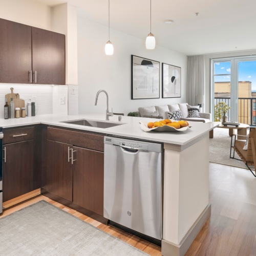 odern kitchen featuring dark wood cabinets, stainless steel appliances, and a white countertop island opening up to a bright living room with hardwood floors and balcony access.